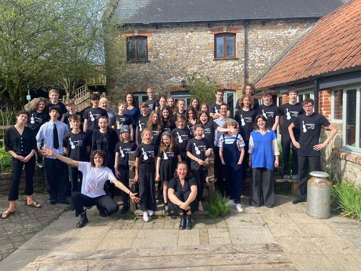 A group of about 30 children and six adults pose together for a group photo. they have their mouths open as if singing and all the children wear a T shirt that says Magdalen Farm Strings on it.