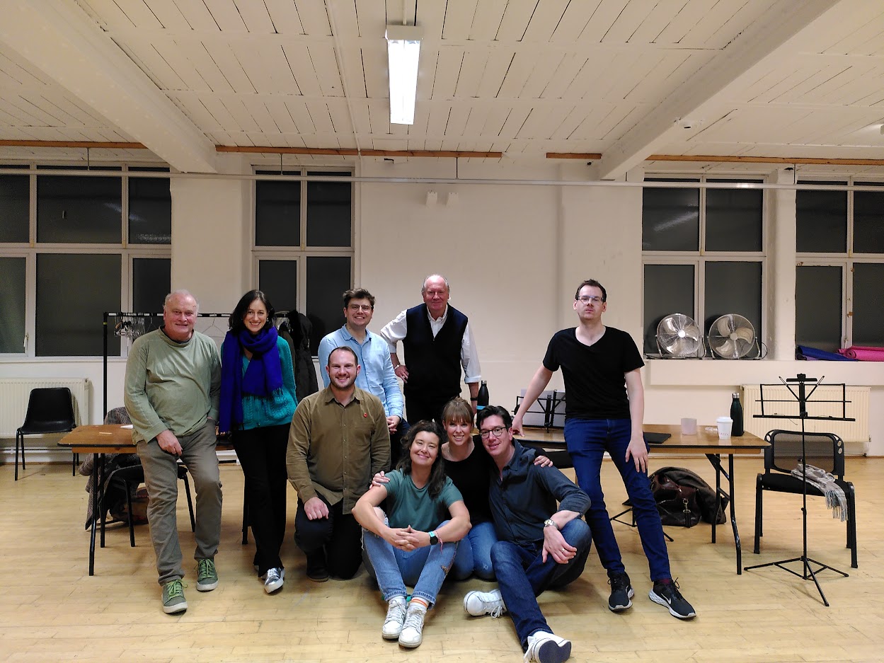 A group of 9 pose together for a photo in a music rehearsal room