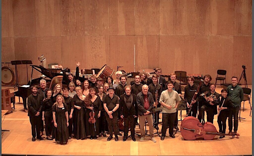 a large group of musicians, some with instruments, stand together on a concert stage